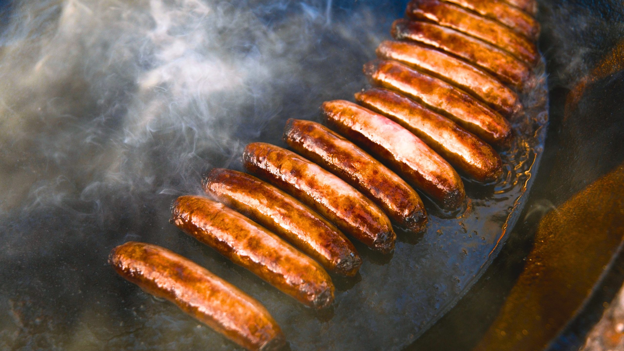 Grilled Chicken Sausage with pork casing sizzling in a pan, surrounded by smoke.