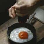 From above of crop anonymous female pouring savory from pepper mill on portion of fried egg in pan