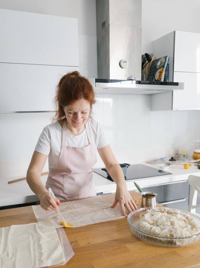 A Woman Wearing Apron while Holding a Pastry Brush and Parchment Paper on the Table