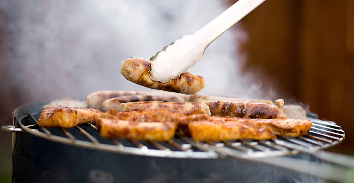 Grilled sausages over an open flame - A close-up view of several grilled sausages cooking over hot grates, with steam and smoke billowing around them.