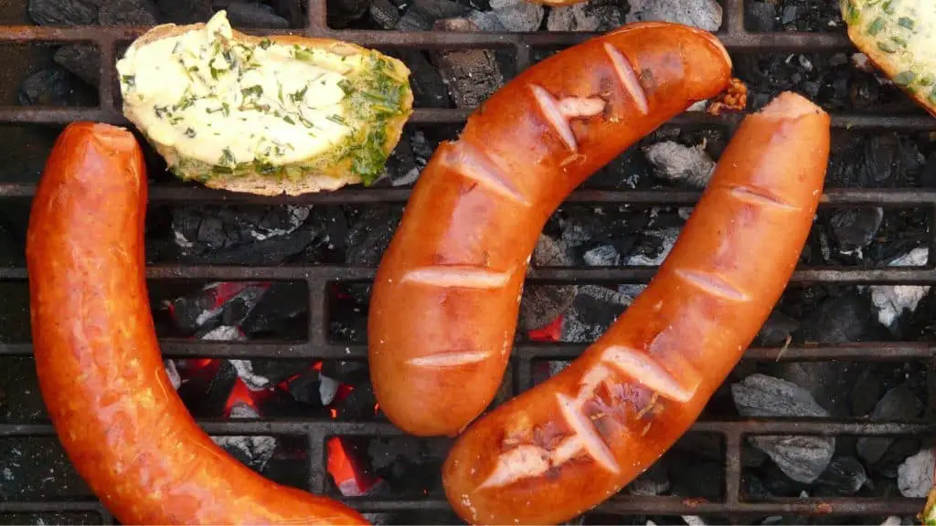 Grilled sausages and slices of bread with herb butter cooking over hot charcoal on a barbecue grill.