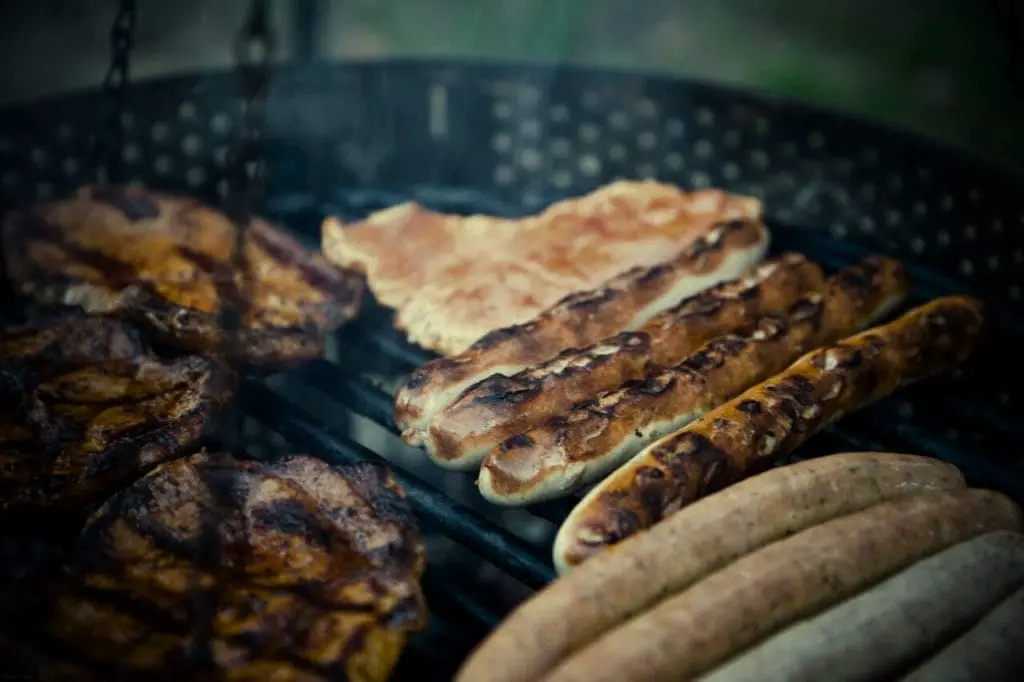 A close-up of sausages and seasoned steaks sizzling on the grates of a smoky barbecue grill.