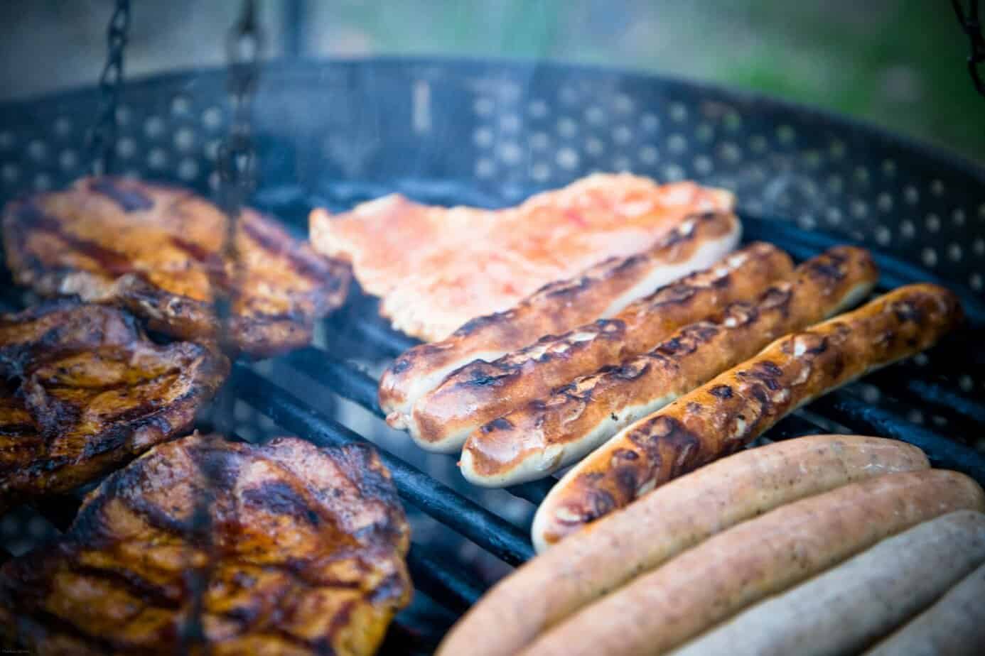 Grilled sausages on a barbecue grill, with various sausages in different stages of cooking, showing charred and golden-brown surfaces.