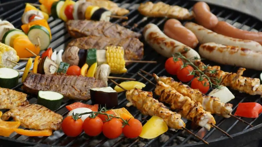Barbecue grill with various grilled meats and vegetables, including kebab skewers, steak, sausages, corn on the cob, bell peppers, zucchini, and cherry tomatoes.