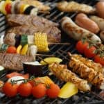 Barbecue grill with various grilled meats and vegetables, including kebab skewers, steak, sausages, corn on the cob, bell peppers, zucchini, and cherry tomatoes.