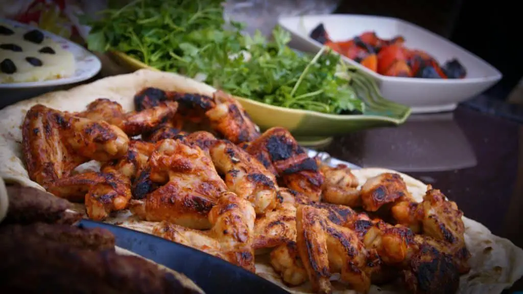 Grilled, caramelized chicken wings with charred edges, served on a flatbread with fresh herbs and roasted vegetables in the background.