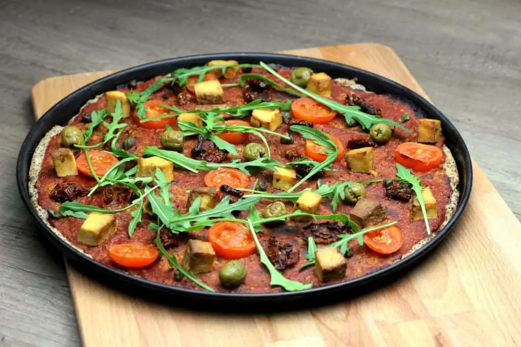 Gluten-free pizza with tomato sauce, cherry tomatoes, green olives, tofu cubes, arugula leaves, and what appears to be sun-dried tomatoes, served on a black plate and wooden cutting board.