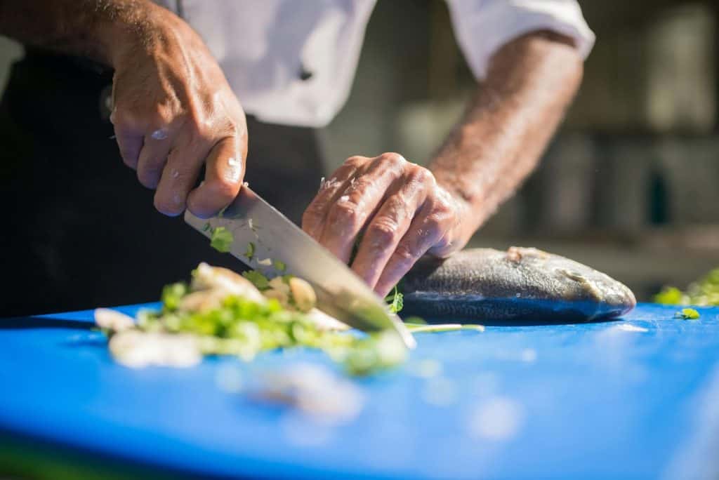 Chef's hands preparing a fish dish with fresh herbs and vegetables on a cutting board.