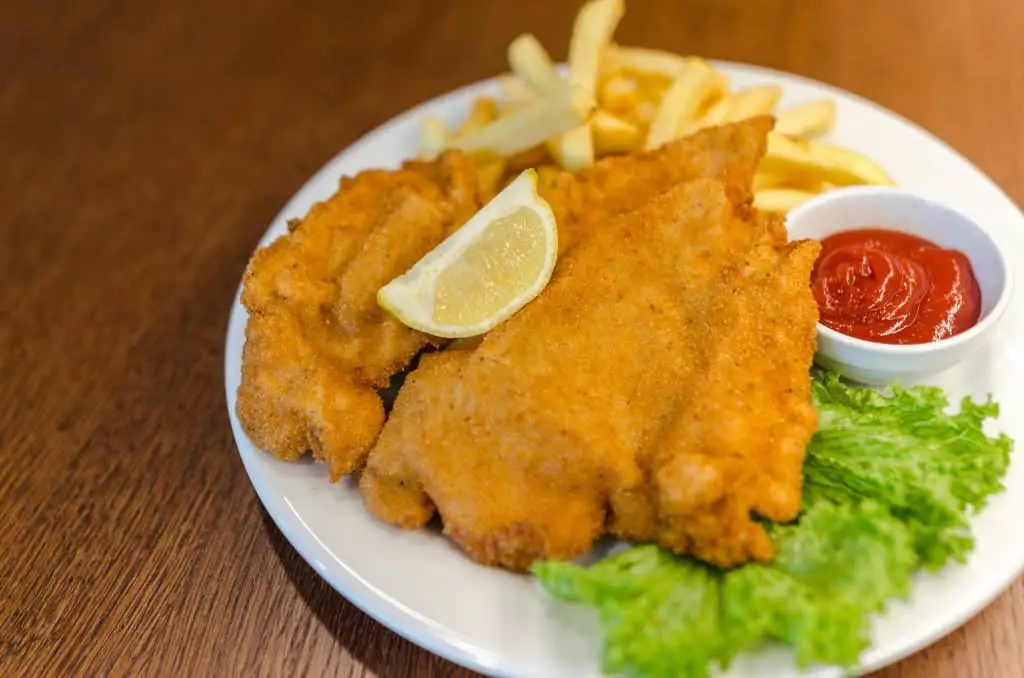 Breaded and fried chicken dish, served with fries, lemon wedge, and condiments on a plate with lettuce garnish.
