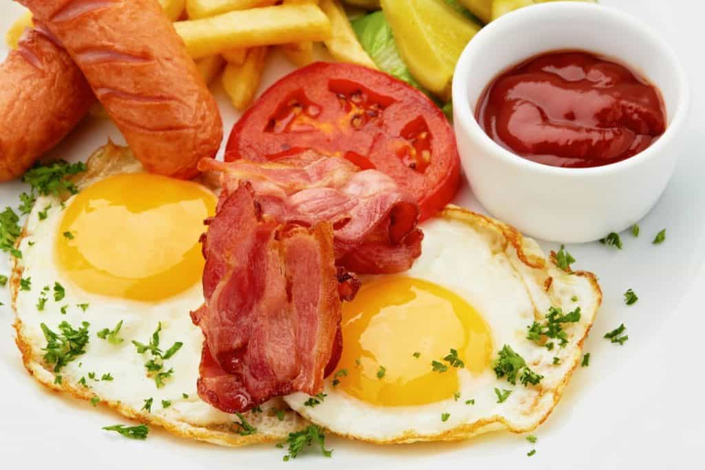 Hearty breakfast plate with sunny-side-up eggs, crispy bacon, sliced tomato, sausages, french fries, and a side of ketchup, garnished with fresh herbs.