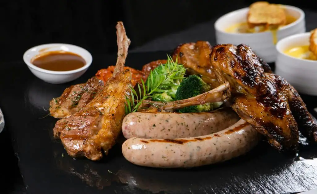 Gourmet plated dish featuring grilled sausages, roasted chicken pieces, broccoli, and herb garnish, served on a black slate with dipping sauces in white bowls.