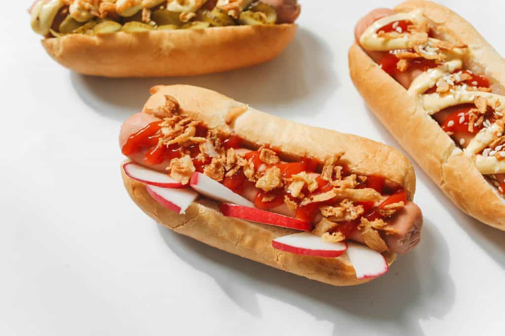 Three gourmet hot dogs with red sauce, crispy toppings, and sliced radishes on a white background, showcasing culinary creativity.