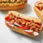 Three gourmet hot dogs with red sauce, crispy toppings, and sliced radishes on a white background, showcasing culinary creativity.