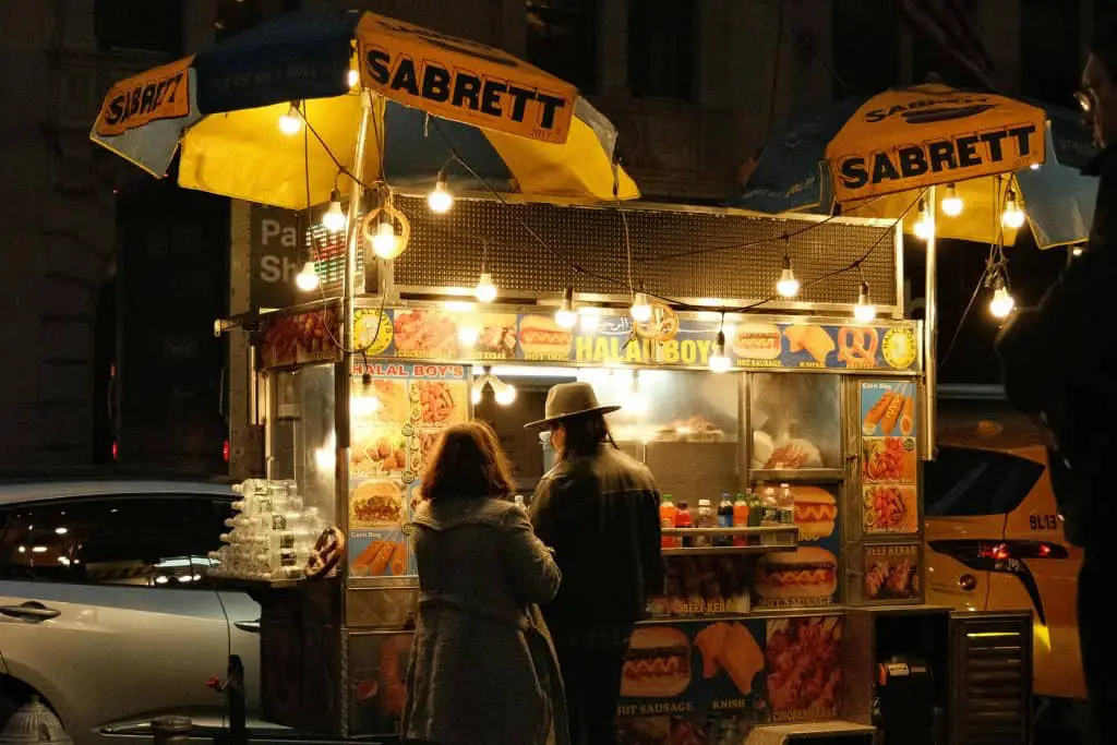 Nighttime street scene of a brightly lit Sabrett hot dog cart with two people ordering, surrounded by colorful menu images and street lights.