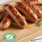 Image shows several grilled sausages on a wooden cutting board, with a gluten-free certification symbol indicating these are gluten-free chicken sausages.